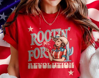 May include: Red t-shirt with the text "Rootin' for Revolution" and a vintage-style illustration of a woman in a cowboy hat holding a sparkler. The design includes stars and a cactus, with a patriotic theme.