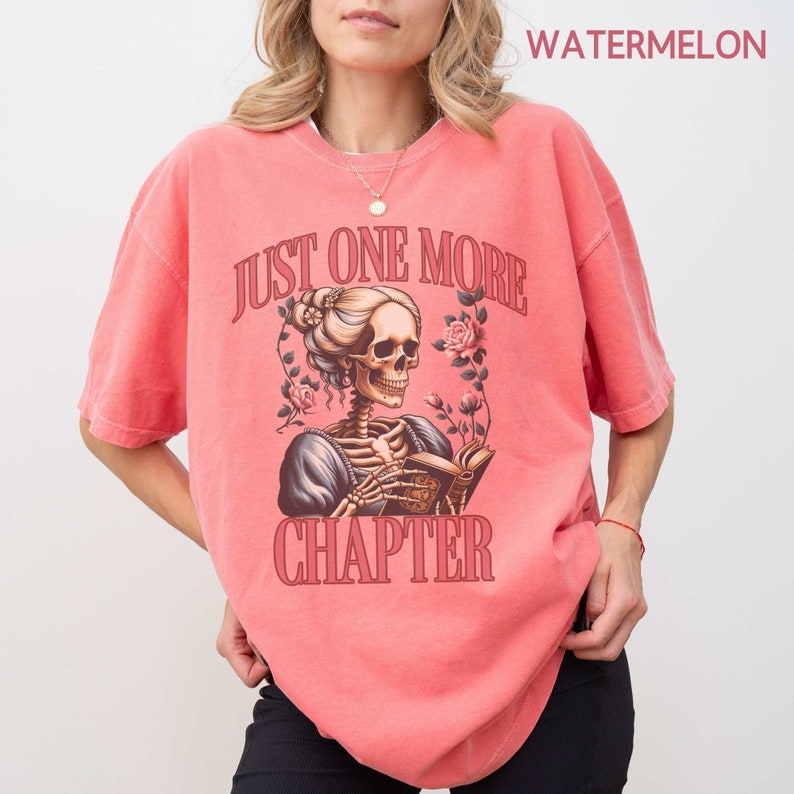 May include: A pink t-shirt with a graphic of a skeleton woman reading a book. The text on the shirt says "Just One More Chapter".