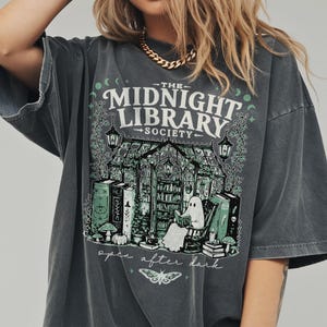 Könnte beinhalten: Ein anthrazitfarbenes Kurzarm-T-Shirt mit einem Grafikdruck eines Gewächshauses, in dem ein Geist ein Buch liest. Der Text lautet THE MIDNIGHT LIBRARY SOCIETY und open after dark. Das Design zeigt Laternen, Bücher, Pilze und einen Nachtfalter.