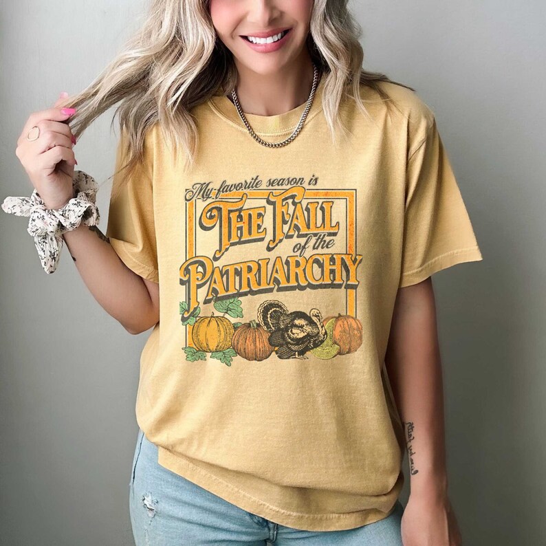 May include: A yellow t-shirt with a vintage-style graphic that says "My favourite season is The Fall of the Patriarchy". The graphic features a turkey and pumpkins.