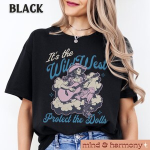 Może przedstawiać: Czarny T-shirt z grafiką kobiety grającej na gitarze przed chmurą z tekstem "It's the Wild West Protect the Dolls".