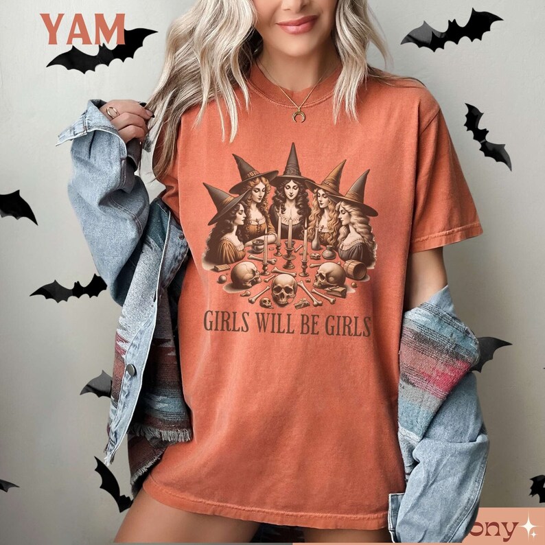Peut inclure: Un t-shirt orange br&ucirc;l&eacute; avec un motif de cinq sorci&egrave;res assises autour d'une table avec des cr&acirc;nes et des os. Le texte "Girls Will Be Girls" est imprim&eacute; sous le motif.