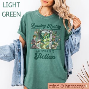 May include: A light green t-shirt with a vintage-style graphic that says "Leaving Reality Stepping Into Fiction". The graphic features a woman reading a book in front of an open doorway leading to a fantastical landscape.