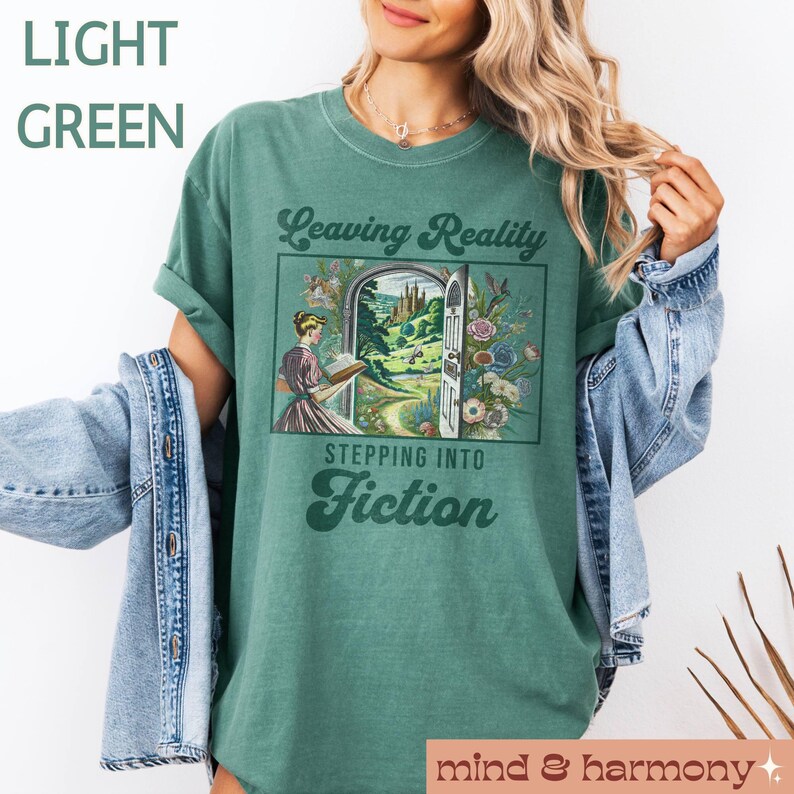 May include: A light green t-shirt with a vintage-style graphic that says "Leaving Reality Stepping Into Fiction". The graphic features a woman reading a book in front of an open doorway leading to a fantastical landscape.
