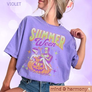 May include: A violet t-shirt with the words "SUMMER Ween" in green and purple. The graphic shows Frankenstein and his bride on a carved pumpkin, holding cocktails. The shirt also has the text "mind & harmony".