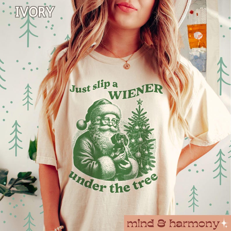 May include: A cream-coloured t-shirt with a green illustration of Santa Claus holding a dachshund under a Christmas tree. The text "Just slip a WIENER under the tree" is printed on the shirt.