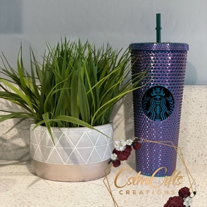 May include: A purple and iridescent reusable cup with a black and white skull graphic and a green lid. The cup is covered in rhinestones and has a straw.