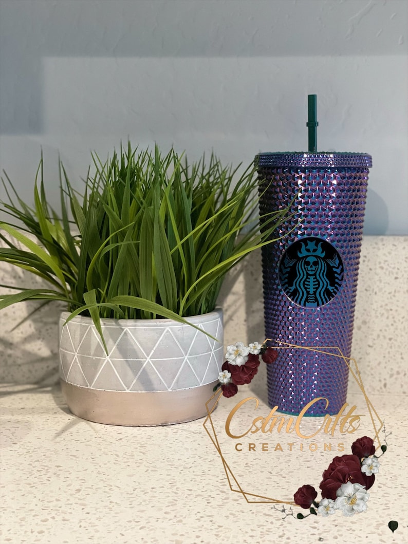 May include: A purple and iridescent reusable cup with a black and white skull graphic and a green lid. The cup is covered in rhinestones and has a straw.