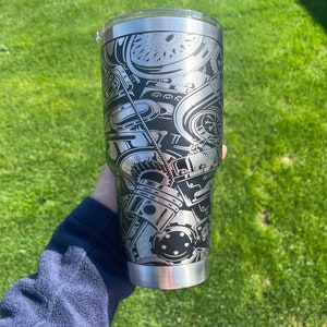 May include: A stainless steel tumbler with a black and silver design of an engine. The tumbler has a lid and is held in a hand.