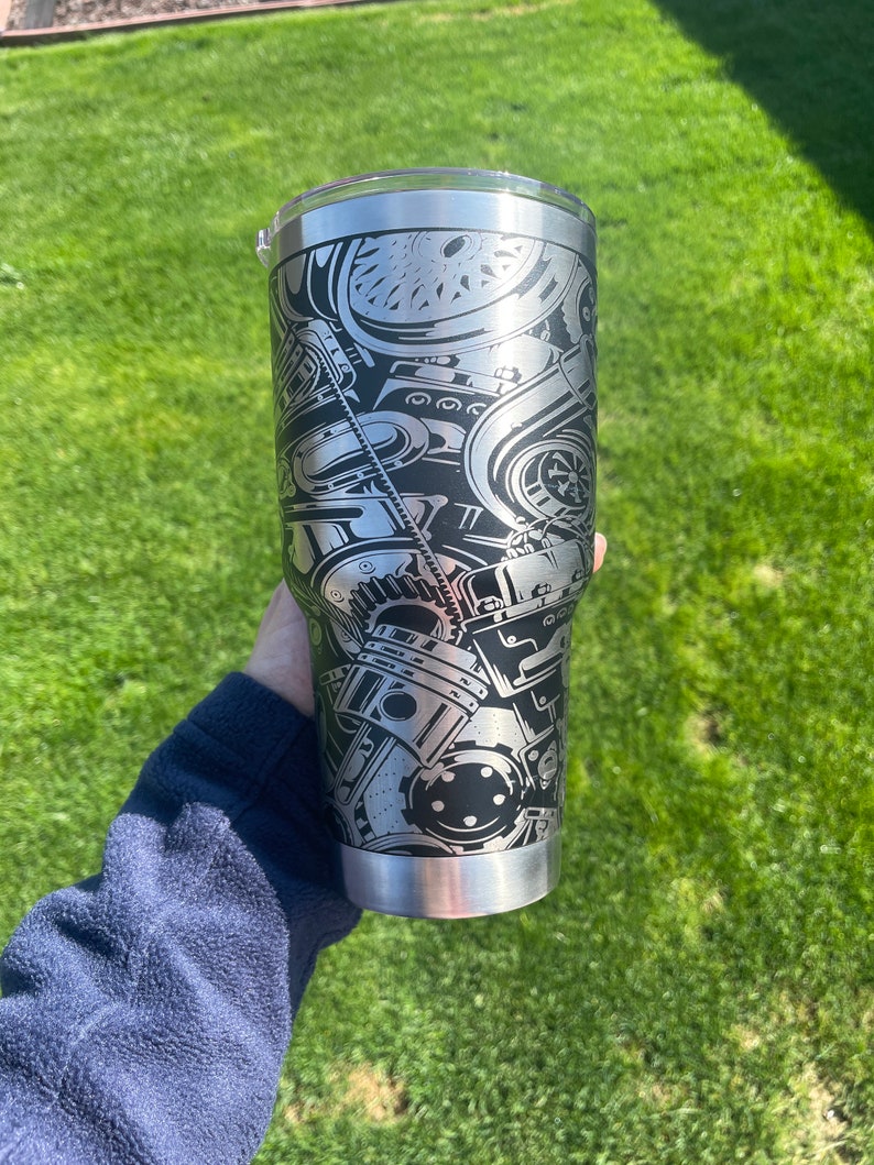 May include: A stainless steel tumbler with a black and silver design of an engine. The tumbler has a lid and is held in a hand.
