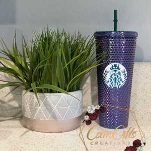 May include: A purple and green iridescent Starbucks cup with a skull and crossbones design. The cup is covered in rhinestones and has a green straw. The cup is sitting on a white countertop with a potted plant in the background.