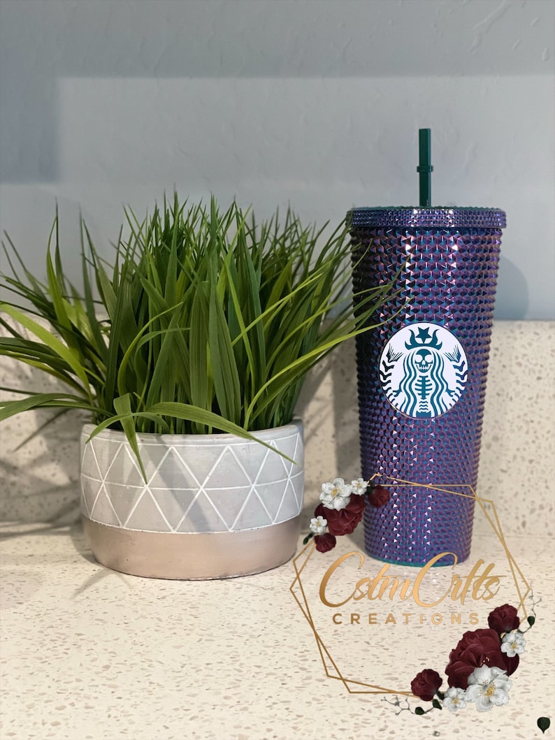 May include: A purple and green iridescent Starbucks cup with a skull and crossbones design. The cup is covered in rhinestones and has a green straw. The cup is sitting on a white countertop with a potted plant in the background.