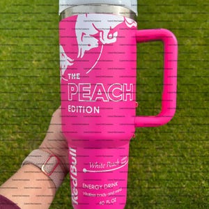 Pu&ograve; includere: Un bicchiere termico rosa con coperchio e manico bianchi. Il bicchiere presenta il testo "THE PEACH EDITION" e "White Peach ENERGY DRINK" con un logo Red Bull. Contiene 1,18 litri.