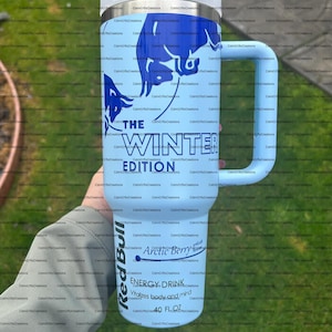 Pu&ograve; includere: Un bicchiere termico azzurro con manico, con una grafica di toro blu e la scritta "THE WINTER EDITION". Mostra anche "Red Bull Arctic Berry" e "ENERGY DRINK 40 FL OZ."