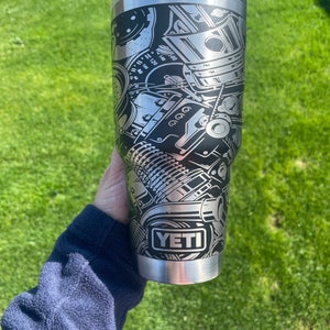 May include: A silver Yeti tumbler with a black and white graphic of a car engine. The tumbler has the word "YETI" printed on the side.
