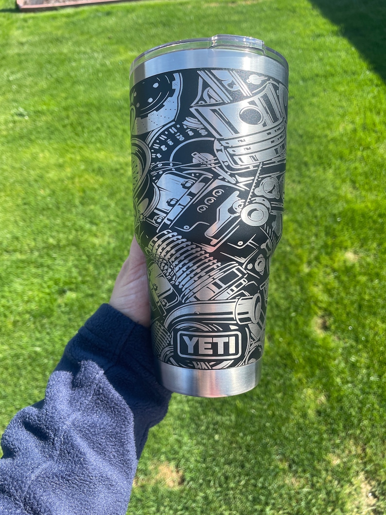 May include: A silver Yeti tumbler with a black and white graphic of a car engine. The tumbler has the word "YETI" printed on the side.
