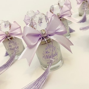 May include: Purple and white favor jars with a crystal-like top and a bow. The jars are decorated with a purple tassel and the text "Miss Nataly 12-3-22".