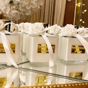 May include: A collection of small, clear glass bottles with white ribbon and a white flower on top. Each bottle has a gold label with the text "Fatima & Ahmed" and "Siz Mutlu" in white lettering.