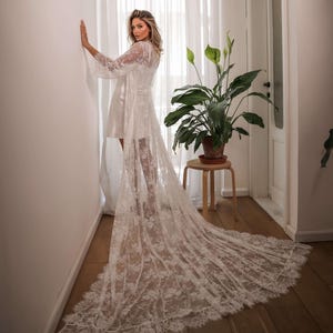 Op de afbeelding: Een witte kanten robe met lange mouwen en een lange sleep. De robe is doorschijnend en heeft een delicaat bloemenpatroon. De robe wordt gedragen door een persoon die in een kamer staat met een plant en een houten kruk.