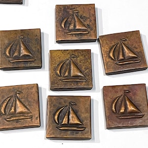 May include: A collection of square, bronze-colored tiles, each featuring a raised sailboat design. The tiles have a weathered appearance, with variations in color and texture. The sailboats have a detailed depiction of the sails and hull.