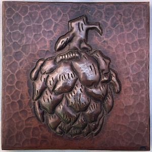 Puede incluir: Una baldosa de cobre con un diseño en relieve de un cono de lúpulo. La baldosa tiene una textura martillada.