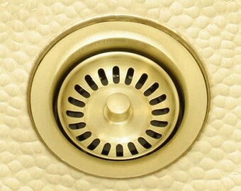 May include: A close-up of a gold-toned kitchen sink strainer with a circular design. The strainer has a series of small holes around the edge and a central button.