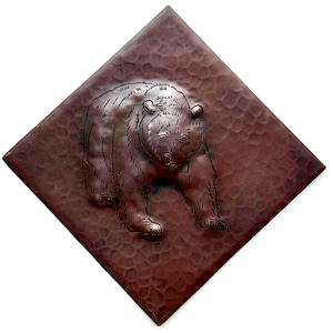 May include: A square, copper-colored wall hanging with a textured surface and a raised relief of a bear. The bear is depicted in a walking pose, with detailed fur and facial features. The overall design is rustic and artistic.