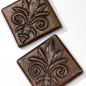 May include: Two square, bronze-colored decorative tiles. Each tile features a raised fleur-de-lis design with intricate details. The fleur-de-lis is centered on the square, with a textured surface and a slightly aged appearance. The tiles are displayed on a white background.