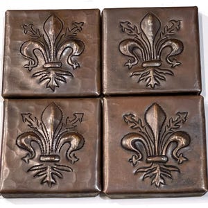 May include: Four square, copper-colored tiles with a hammered texture. Each tile features a raised fleur-de-lis design in the center. The tiles are arranged in a two-by-two grid, showcasing the repeating pattern and metallic finish.