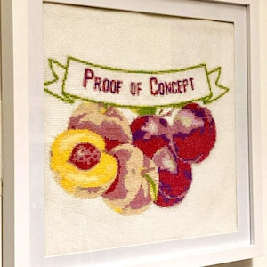 May include: Framed cross-stitch artwork featuring a cluster of fruits, including a sliced peach, and plums in shades of red, purple, and yellow. A green banner above the fruits reads "PROOF OF CONCEPT" in purple letters. The artwork is framed in a white wooden frame.