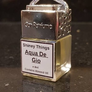 May include: A silver and glass car air freshener with a black label that reads "Shiny Things Aqua De Gio e 8ml Contains Almond Oil".