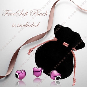 Puede incluir: Tres abalorios en forma de coraz&oacute;n rosa con detalles plateados, dispuestos sobre una superficie blanca con una bolsa de terciopelo negro y una cinta rosa. El texto "Free Soft Pouch is included" est&aacute; impreso en la superficie blanca.
