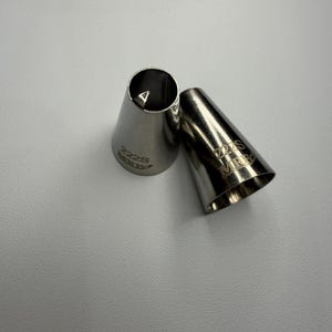 Könnte beinhalten: Zwei Spritztüllen aus Edelstahl. Eine Tülle steht aufrecht und zeigt eine sternförmige Öffnung, die andere liegt auf der Seite. Auf der aufrechten Tülle sind die Nummer "222S" und das Wort "MERCK" eingraviert.
