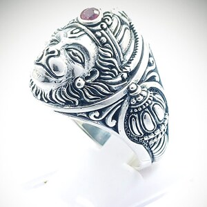 Op de afbeelding: Een zilveren ring met een sierlijk ontwerp met een gezicht en een rode edelsteen. De ring is versierd met ingewikkelde gravures en een kroon.