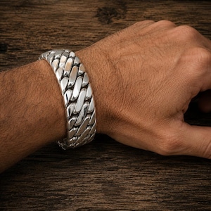 May include: A wide, silver-colored chain bracelet with a polished finish. The bracelet is made of interlocking links and is worn on a wrist. The background is a dark wood surface.