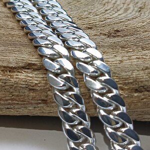 May include: Close-up of two silver curb chains. The chains are made of interlocking, rounded links that reflect light. The chains are placed on a light brown wooden surface, creating a contrast between the shiny metal and the natural wood.