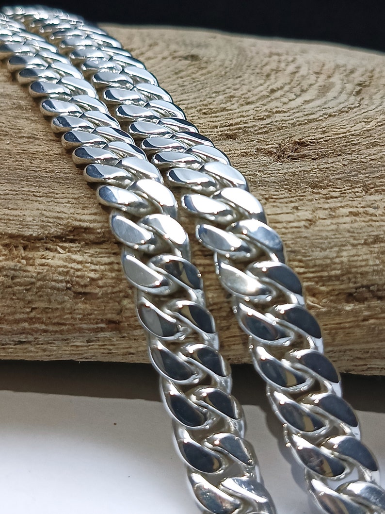 May include: Close-up of two silver curb chains. The chains are made of interlocking, rounded links that reflect light. The chains are placed on a light brown wooden surface, creating a contrast between the shiny metal and the natural wood.