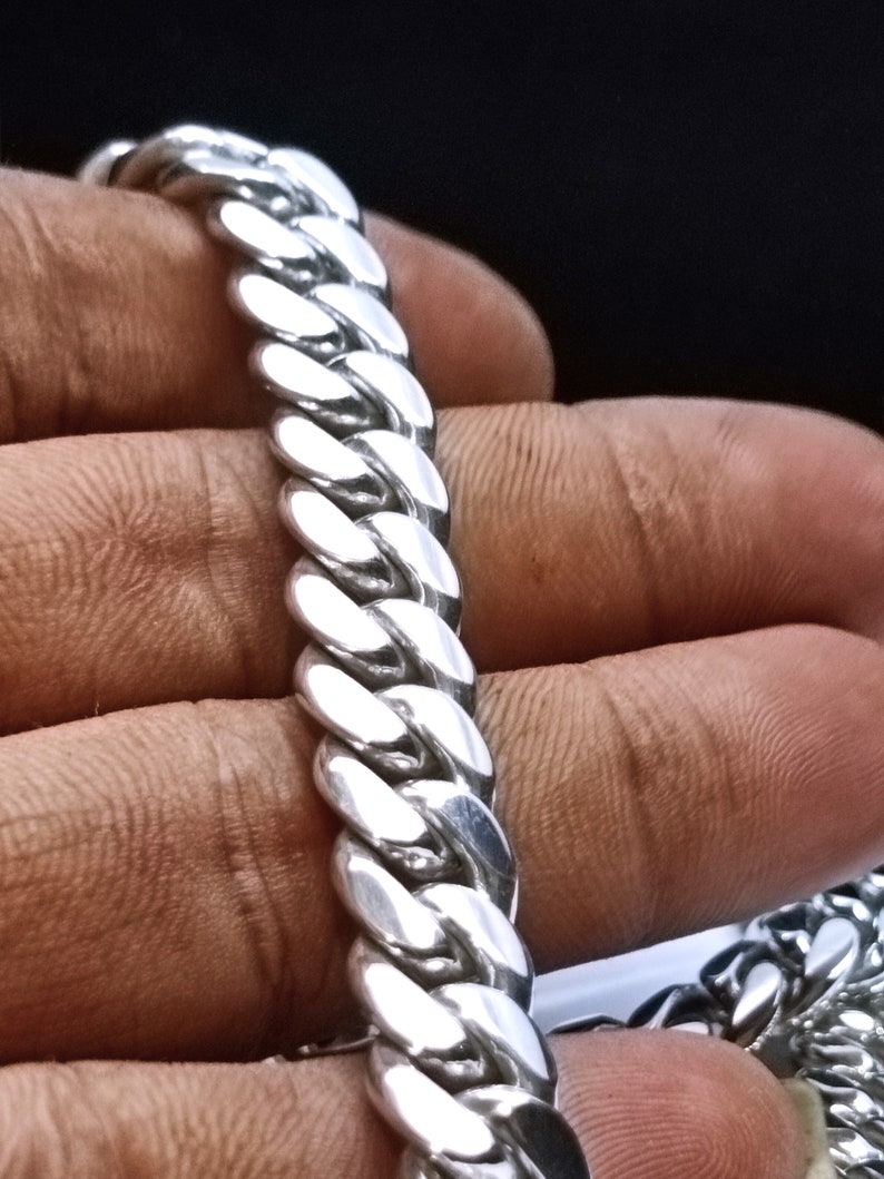 May include: Close-up of a silver curb chain bracelet. The links are thick and oval-shaped, creating a bold, textured appearance. The bracelet is held against a dark background, highlighting its polished, reflective surface. A piece of jewellery.