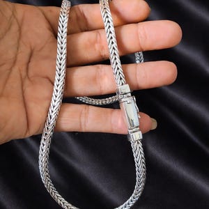 May include: A silver-colored necklace with a woven chain design. The necklace is held against a black background, showcasing its intricate pattern and a secure clasp.