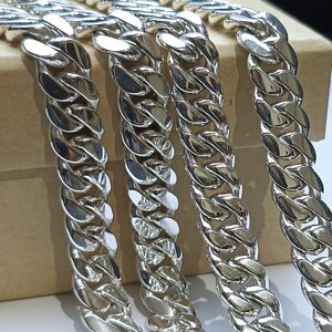 May include: Close-up of several silver curb chains. The chains are thick and have a polished, reflective surface. The links are interlocked, creating a bold design. The chains are displayed on a neutral background.