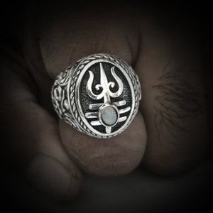 May include: A silver ring with an ornate design featuring a trident symbol and a white opal stone in the centre.