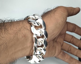 May include: A silver-colored curb chain bracelet. The bracelet is made of thick, interlocking links. The shiny metal reflects light, creating highlights and shadows. The bracelet is worn on a wrist.