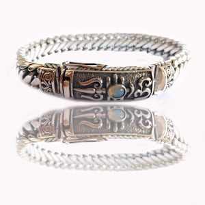 Puede incluir: Una pulsera de plata con un cierre detallado que presenta un símbolo Om de plata y una piedra de ópalo azul. La pulsera tiene un diseño de cadena trenzada.