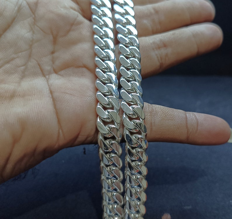 May include: Two silver curb chains with a polished finish. The chains are made of interlocking links, creating a bold and classic design. The links are thick and reflect light. The chains are displayed against a neutral background.