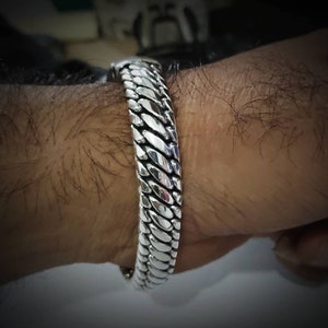 May include: A silver bracelet with a textured, scale-like design. The bracelet is worn on a wrist, showcasing its intricate pattern and polished finish. The bracelet appears to be made of metal and has a secure clasp.
