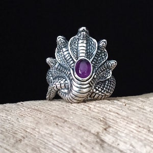 Op de afbeelding: Een zilveren ring met een ontwerp van een slang met drie koppen en een paarse edelsteen in het midden.