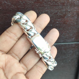 May include: A silver bracelet with a curb chain design. The bracelet has a rectangular plate in the centre. The bracelet is held in a hand.