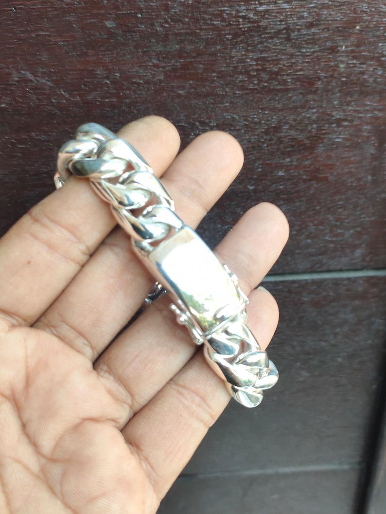 May include: A silver bracelet with a curb chain design. The bracelet has a rectangular plate in the centre. The bracelet is held in a hand.