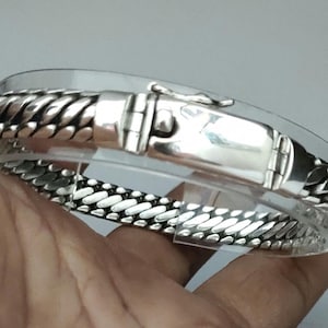 May include: A silver bracelet with a braided design and a clasp. The bracelet is on a hand.