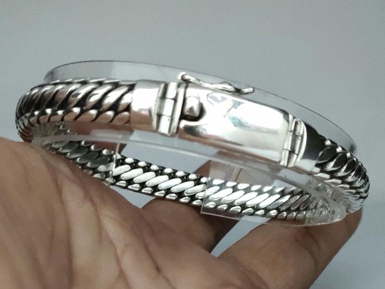 May include: A silver bracelet with a braided design and a clasp. The bracelet is on a hand.